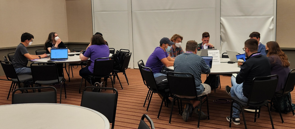 Image showing a room with round tables and people working at each table most of who have laptops sitting in front of them.' style='width: 100%;